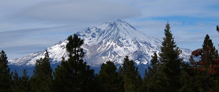 [Behind and above a screen of evergreens is a tall, mostly snow-covered mountain. Some strips of light grey rock are also visible. The sky is mostly white clouds although there is a peek of blue.]